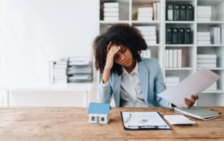 Stressed woman in light blue blazer holding papers at desk with model house, calculator, and clipboard, symbolizing foreclosure concerns